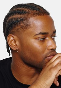 Primo piano di un giovane con capelli intrecciati, che indossa una maglietta nera. Le caratteristiche includono un orecchino e una pelle liscia con toni naturali.