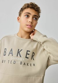 Sudadera beige de manga larga, con cuello redondo y texto impreso "BAKER LONDON" en letras negritas negras en el pecho. Textura suave.