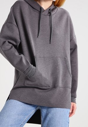 Osoba ubrana w luźną, ciemnoszarą bluzę z kapturem z przednią kieszenią i sznurkami, w połączeniu z jasnoniebieskimi dżinsami na jednolitym tle.