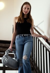 Top preto sem mangas, jeans rasgados azuis claros e uma bolsa cinza larga, de pé nas escadas com corrimãos de metal em um espaço bem iluminado.