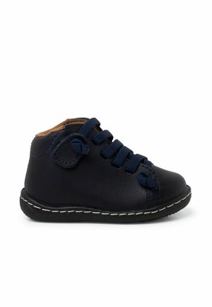 Chaussure en cuir noir avec des lacets bleu marine et une semelle blanche cousue. Dispose d'un bout arrondi et d'un revêtement doux, couleur beige, à la cheville.