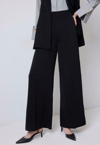 Black wide-leg pants made of smooth fabric with a high waist and subtle front crease, paired with pointed black heels.