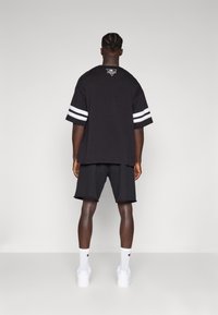 T-shirt de algodão preto com mangas às riscas brancas, apresentando um detalhe de logótipo nas costas. Combinado com calções pretos e tênis brancos.