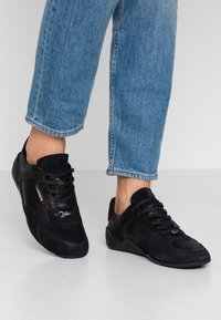 Chaussures de sport noires alliant suède et matériaux texturés, bout arrondi, fermeture à lacets, et petit détail logo sur le côté.