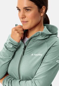 Giacca con cappuccio verde, strutturata e con zip frontale, con il logo "VAUDE" in bianco sul petto e sul maniche sinistra.