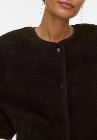 Sieviete tērpusies tumši brūnā faktūrētā vilnas jaku ar apaļu izgriezumu un melniem sprādzīšu pogām, redzamas garas krītošas auskaru pāri.