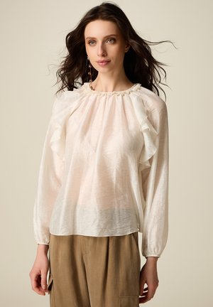 Nainen, joka pukeutunut läpikuultavaan valkoiseen pitkähihainen röyhelöpaitaan ja vaaleanruskeisiin korkeavyötäröisiin housuihin neutraalia taustaa vasten.