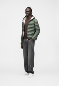 Giacca a piumino verde con motivo trapuntato orizzontale, interno bianco, chiusura con cerniera, indossata sopra un maglione marrone e jeans grigi. Scarpe da ginnastica bianche.