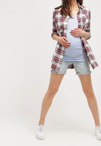 Schwangerschaftsoutfit mit einem hellblauen Oberteil, einem karierten Hemd und Jeansshorts. Das karierte Hemd hat Rottöne und Weiß und ist mit hochgekrempelten Ärmeln gestaltet.