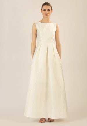 Woman with pulled-back hair wearing a sleeveless white textured floor-length gown and silver open-toe heels against a plain background.