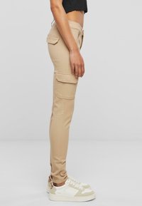 Béžové cargo nohavice so slim fit strihom, bočnými vreckami a jemnou tkaninou, skombinované s bielymi teniskami s béžovými akcentmi.