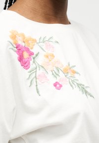 Λευκό ένδυμα με κεντημένα ροζ, ροδακινί και πορτοκαλί λουλούδια και πράσινα φύλλα στο πάνω μέρος του στήθους κοντά στον λαιμό.