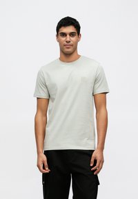 Ανοιχτό γκρι βαμβακερό μπλουζάκι με στρογγυλή λαιμόκοψη και κοντά μανίκια, με μικρό τετράγωνο λογότυπο στο στήθος, συνδυασμένο με μαύρο παντελόνι.