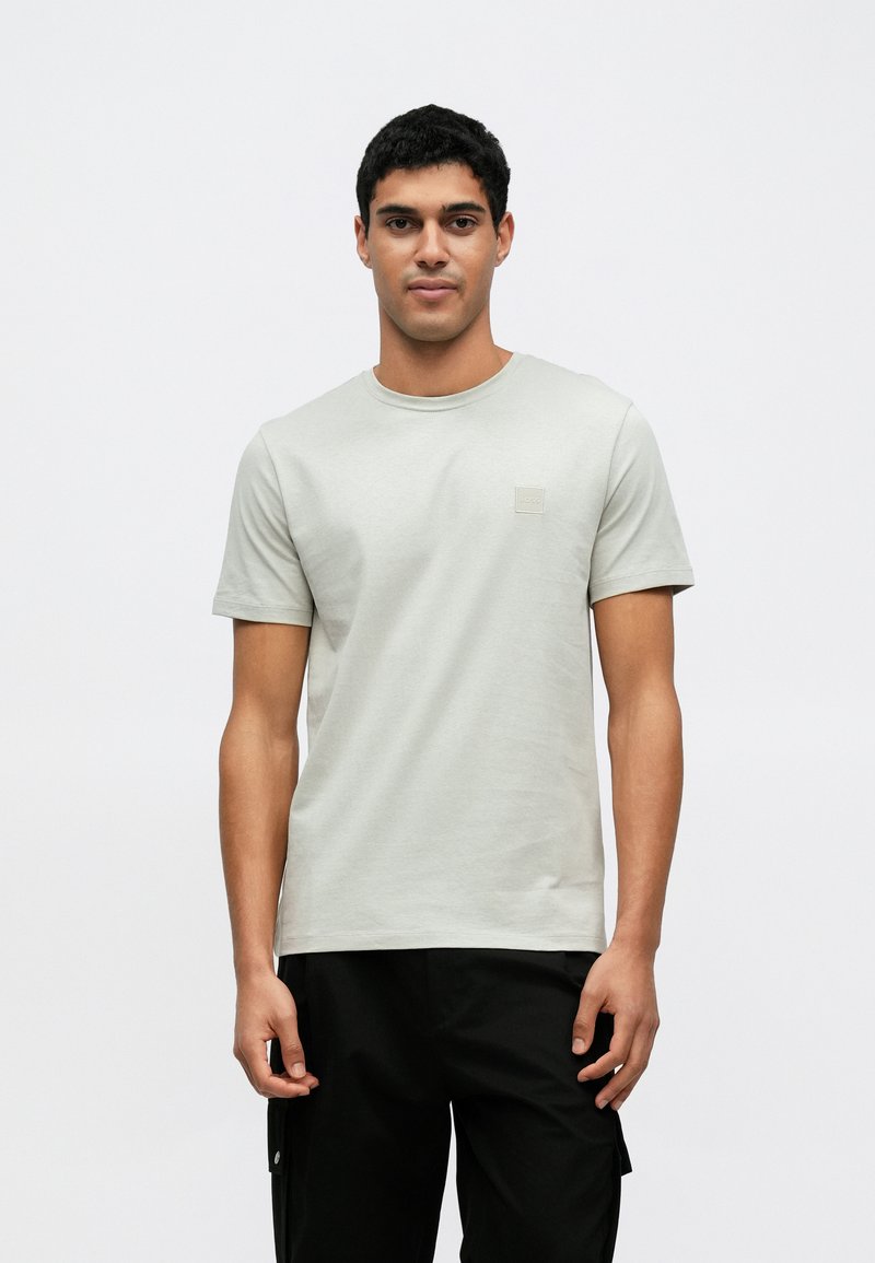 Ανοιχτό γκρι βαμβακερό μπλουζάκι με στρογγυλή λαιμόκοψη και κοντά μανίκια, με μικρό τετράγωνο λογότυπο στο στήθος, συνδυασμένο με μαύρο παντελόνι.