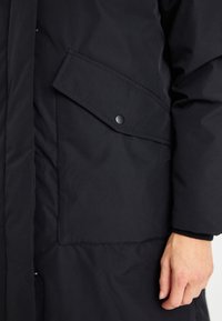 Crna izolirana parka s glatkom površinom, koja ima bočni džep s dugmetima na zatvaranju i visoki ovratnik za dodatnu toplinu.