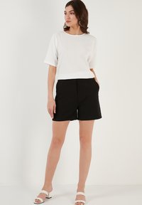 Witte, textuurde top met korte mouwen gecombineerd met zwarte shorts. Opvallende details zijn de zakken op de shorts en de blokhak sandalen.