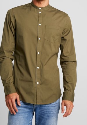 Man in een olijfgroene overhemd met mandarin kraag en borstzak, gecombineerd met lichtblauwe jeans.
