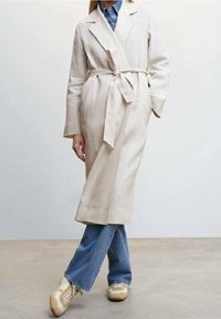 Beige linned trenchcoat med bælte i taljen, åben front og lange ærmer, parret med blå flared jeans og hvide sneakers.