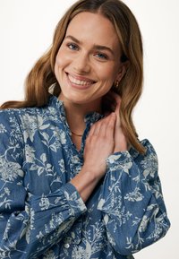 Modrá blůza s květinovým vzorem vyrobená z měkké, texturované látky. Má volánkový výstřih a manžety na knoflíky. Dlouhé rukávy, uvolněný střih.