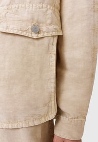 Chaqueta beige con un tejido texturizado, gran bolsillo frontal con cierre de botón plateado y costuras visibles a lo largo de los bordes.