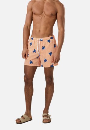 Man zonder shirt die oranje zwemshorts draagt met blauwe schildpaddruk en beige sandalen, staand tegen een witte achtergrond.