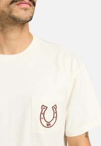 White cotton t-shirt with short sleeves, featuring a front pocket. Embroidered horseshoe design in maroon with "VA" underneath.