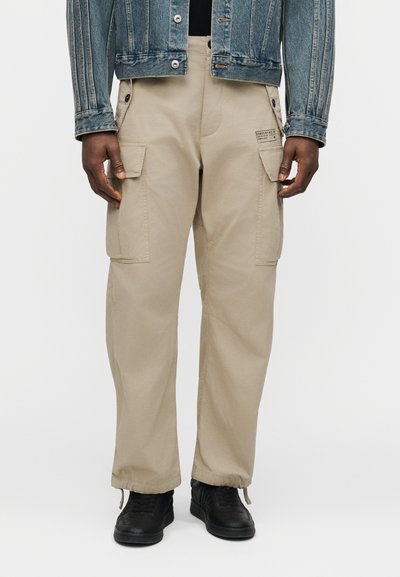 Homme portant un pantalon cargo beige avec des poches à boutons et des ourlets à cordon de serrage, associé à une veste en denim et des baskets noires.