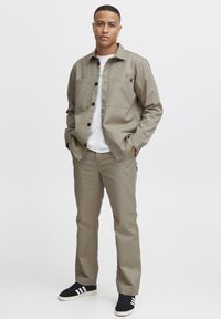 Light khaki jacket with button closure and two front pockets, paired with matching trousers and black sneakers with white stripes.