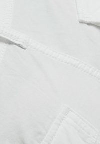 Vitt bomullstyg med en mjuk textur, med en synlig ficka och sömda detaljer. Designen är enkel och odekorerad.