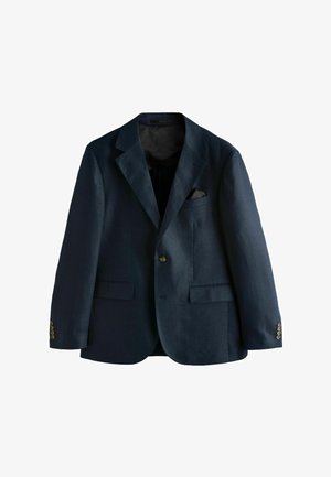 Muž upravuje gombík na rukáve tmavomodrého obleku, vidieť je biely manžeť košele a čiastočne zobrazená vzorovaná kravata.