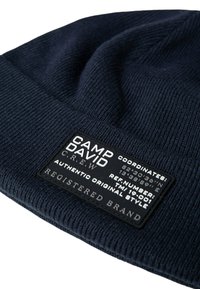 Navy blue knitted beanie with textured fabric, featuring a black label with white text displaying brand name and coordinates.