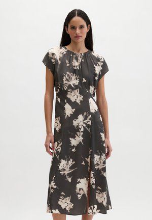 Vrouw met lang donker haar, gekleed in een zwarte en beige midi-jurk met bloemenprint, korte mouwen en een split vooraan, staande tegen een effen achtergrond.