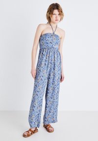 Modra bezramna kombinezona s cvetnim paisley vzorcem, z elastičnim pasom in širokimi hlačnicami. Model nosi rjave sandale na naramnice.
