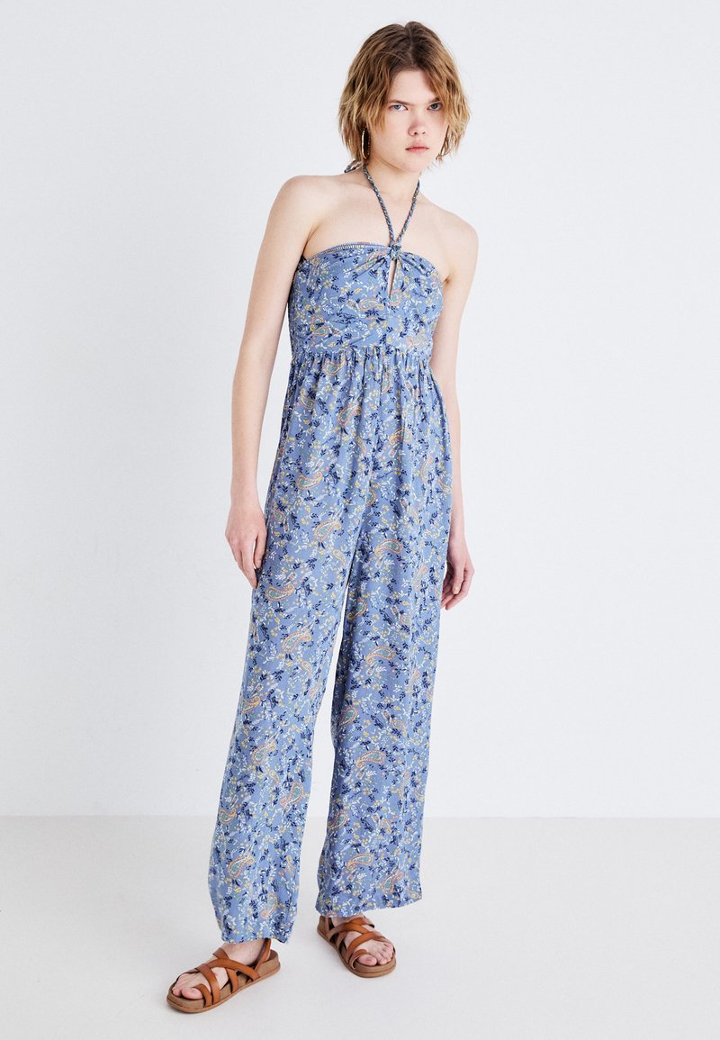 Modra bezramna kombinezona s cvetnim paisley vzorcem, z elastičnim pasom in širokimi hlačnicami. Model nosi rjave sandale na naramnice.