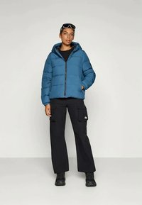 Casaco acolchoado em teal com capuz, fecho de correr e detalhes acolchoados. Combinado com calças cargo pretas e botas pretas, colocado contra um fundo neutro.