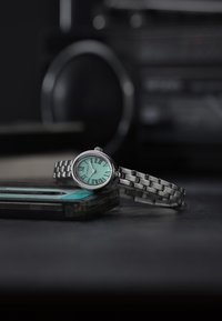 Hõbedane metallist kell ovaalse näo, münti rohelise taustaga ja rooma numbritega, millel on seotud käevõru disain. Asetatud kassettide peale.