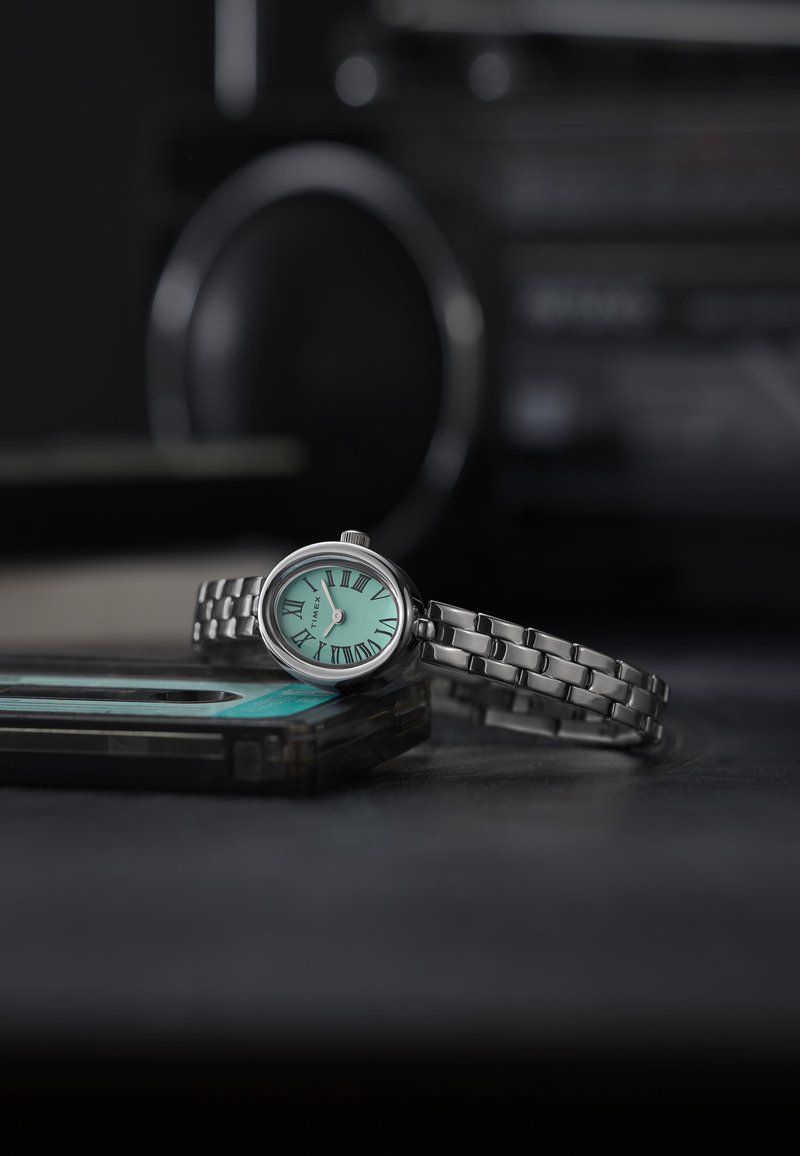 Hõbedane metallist kell ovaalse näo, münti rohelise taustaga ja rooma numbritega, millel on seotud käevõru disain. Asetatud kassettide peale.