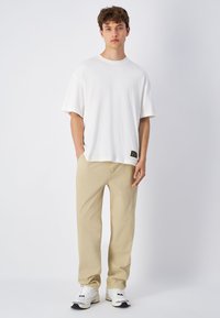 T-shirt bianca a manica corta con una vestibilità rilassata, abbinata a pantaloni beige. L'outfit è completato da sneakers bianche con dettagli neri.