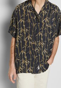 Camicia a maniche corte in tessuto blu nave con stampa di bambù in verde e beige. Ha una chiusura con bottoni e una vestibilità morbida.
