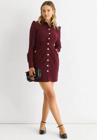 Woman wearing a burgundy button-up dress with long sleeves, holding a black clutch, and wearing black ankle-strap heels against a white background.