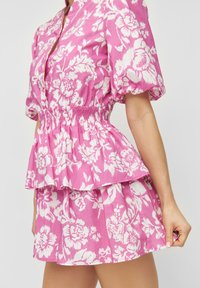 Žena v růžovém květovaném dvoudílném oblečení s nařasenými krátkými rukávy a elastickým pasem, která drží lem sukně.