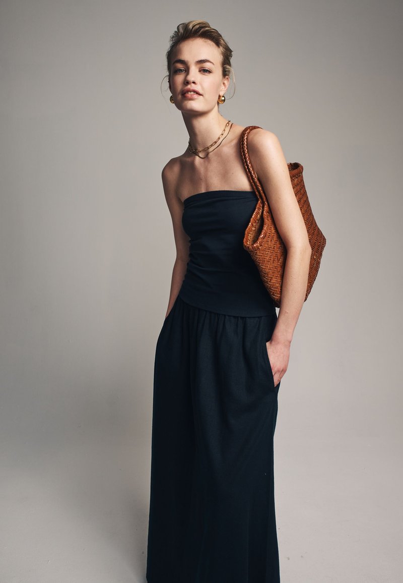 Zwart strapless top met een aansluitend lijfje, gecombineerd met losse zwarte broek met wijde pijpen. Model draagt een gestructureerde bruine geweven tas.
