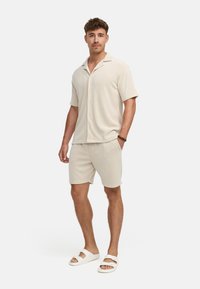 Beige Kurzarmhemd mit Knopfverschluss, kombiniert mit passenden Shorts. Weicher gerippter Stoff. Getragen mit weißen Slipper-Sandalen.