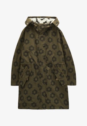 Kobieta nosząca zielony płaszcz z kapturem w kwiatowy wzór, ciemnoniebieskie spodnie, brązowe buty z zielonymi sznurowadłami, trzymająca białą torbę z ciemnym kwiatowym nadrukiem.