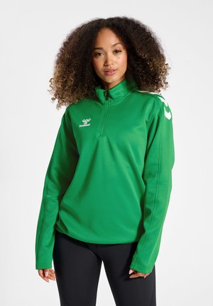 Kvinde med krøllet hår iført en grøn sportstop med kvart-lynlås med hvidt logo og sorte bukser, stående mod en hvid baggrund.