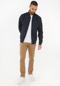 Giacca zip-up blu navy con colletto alto, due tasche frontali, caratterizzata da una finitura liscia. Abbinata a pantaloni chinos beige e sneaker bianche.