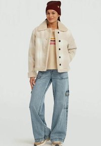 Beige fleece jacket with a shearling collar, paired with a beige shirt featuring stripes and wide blue denim cargo pants.
