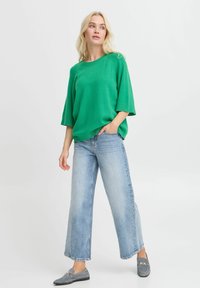 Grüner, übergroßer Strickpullover mit kurzen Ärmeln, kombiniert mit hellblauen, weit geschnittenen Jeans und grauen Slippern, stehend vor einem neutralen Hintergrund.
