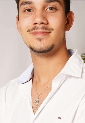 Junger Mann mit gepflegtem Gesichtshaar, der ein weißes Hemd mit Kragen und eine silberne Kreuzkette trägt und leicht lächelt vor einem neutralen Hintergrund.