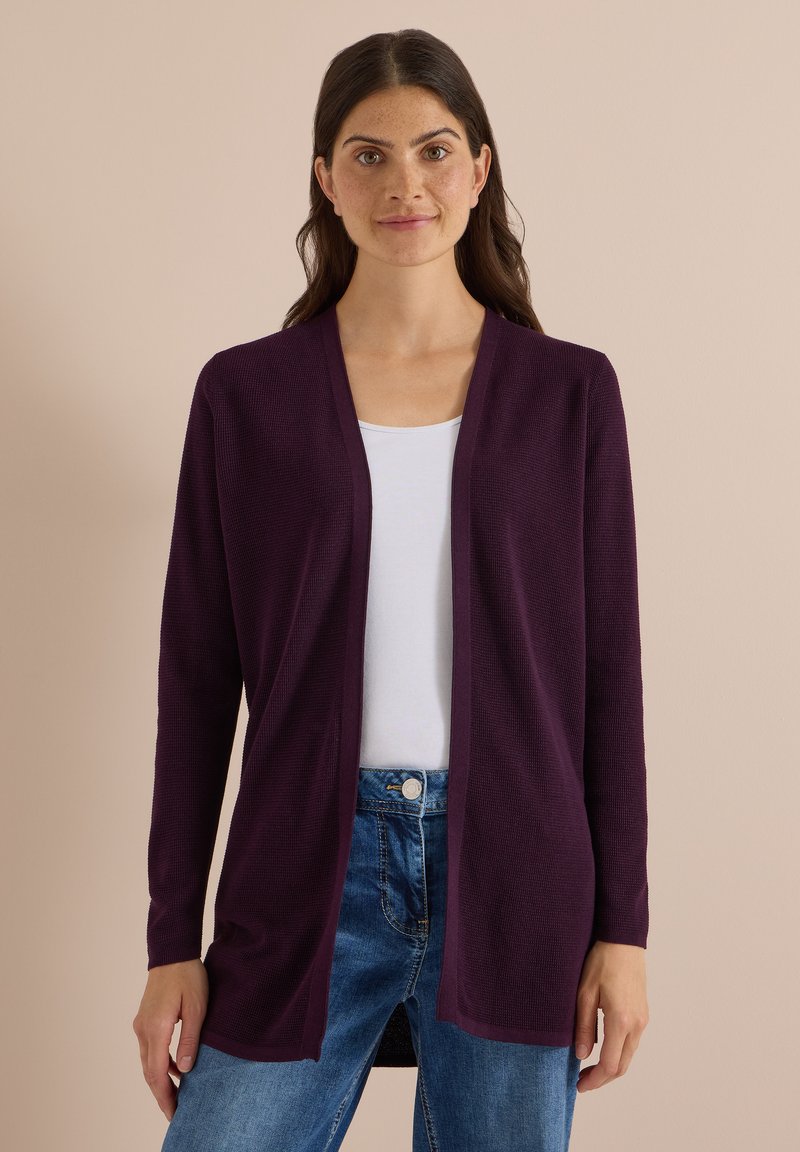 Femme aux cheveux foncés portant un long cardigan violet ouvert sur un haut blanc et un jean bleu sur un fond beige uni.